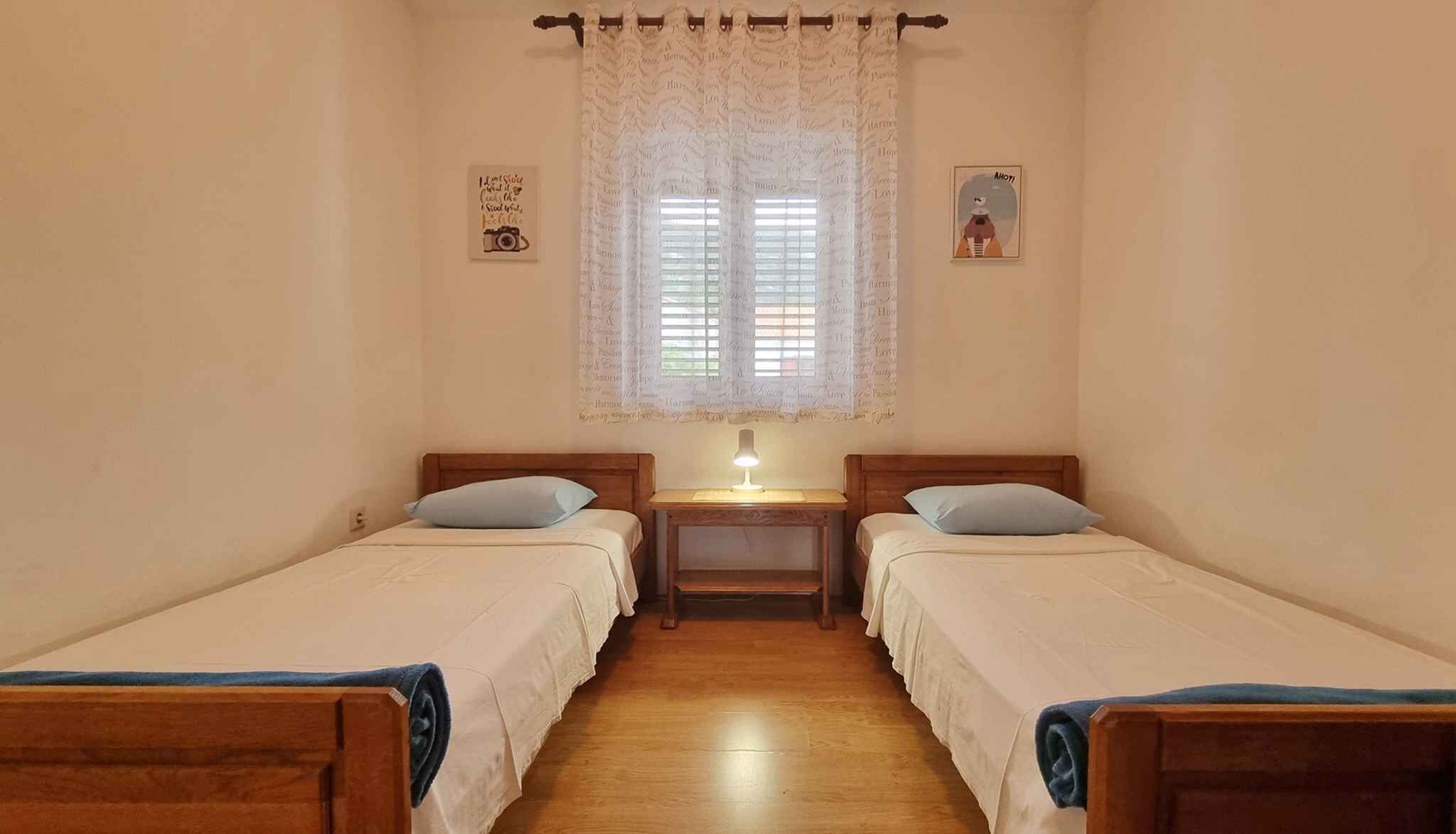 Una habitación pequeña y acogedora con dos camas individuales, una mesita de noche con lámpara, una ventana con cortinas de encaje y un cuadro en la pared.