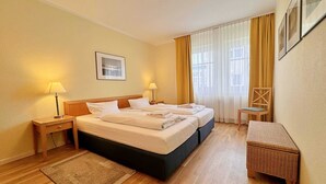 Une chambre d’hôtel avec un lit double, deux tables de chevet avec des lampes, une petite chaise en bois, un banc en osier et une grande fenêtre avec des rideaux.