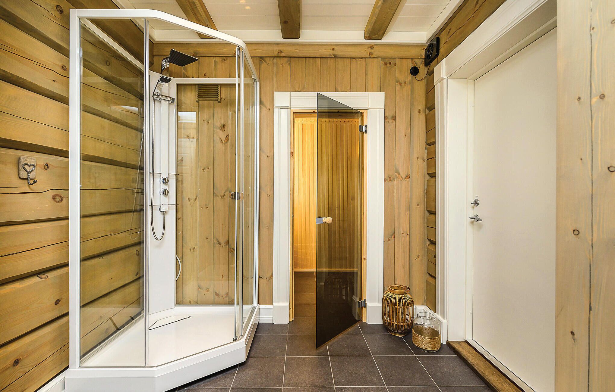A wooden interior with a glass-enclosed shower, a door leading to another room, and a tiled floor.