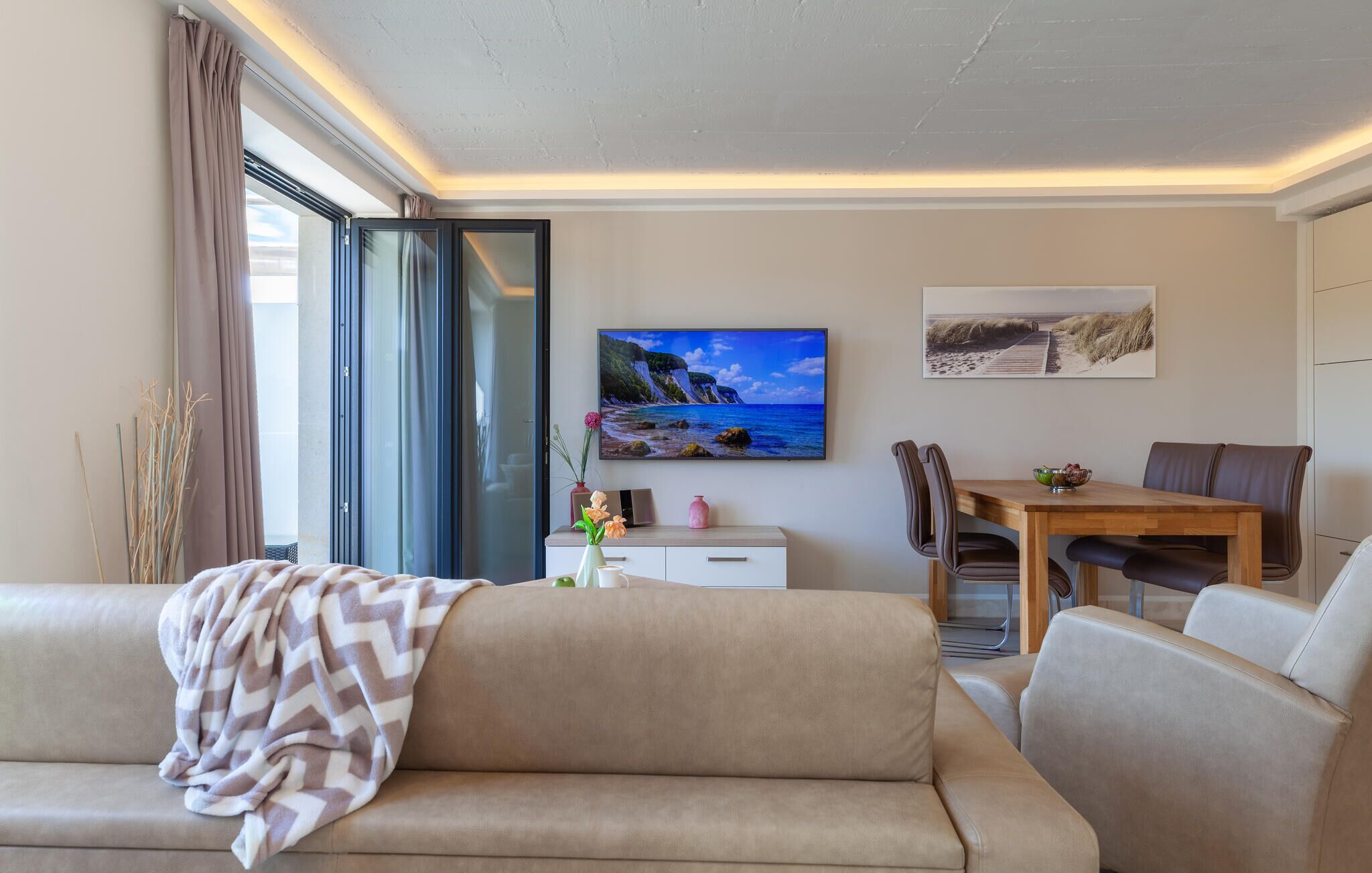 A modern living room with a sofa, a dining table with chairs, a TV, and artwork on the wall.