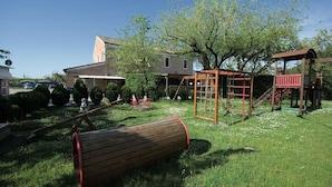 Un terrain de jeux avec des balançoires, un toboggan et une structure d’escalade, situé dans un espace verdoyant avec des arbres et une maison en arrière-plan.