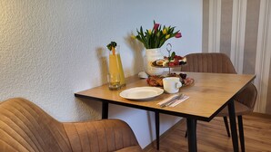 Una mesa de comedor con sillas, un florero con flores, una cesta de frutas, un croissant, una taza y un vaso de jugo.