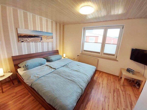 A bedroom with a wooden bed, a nightstand, a bedside table, a TV, and a window with blinds.