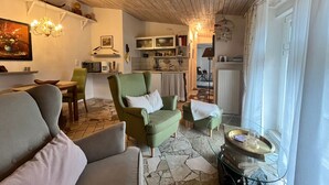 A cozy living room with a green armchair, a sofa, and a dining table. There is a kitchen area with cabinets and a window. - Midsummer Night's Dream - Kunsthof Salsitz (Lohme auf Rügen)