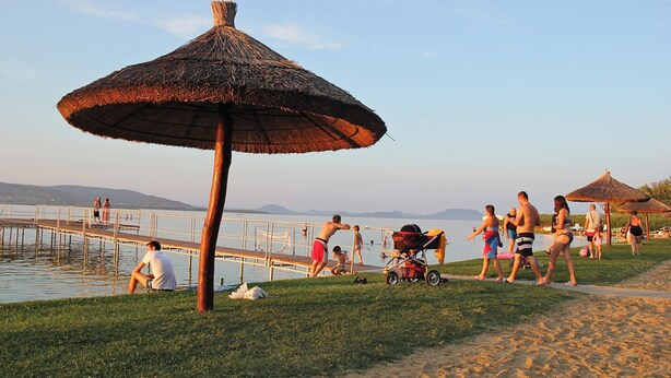 A beach scene with a thatched umbrella, a pier, and people walking along the shore. - Ferienhaus mit Außenpool und großem Garten (Balatonkeresztúr)