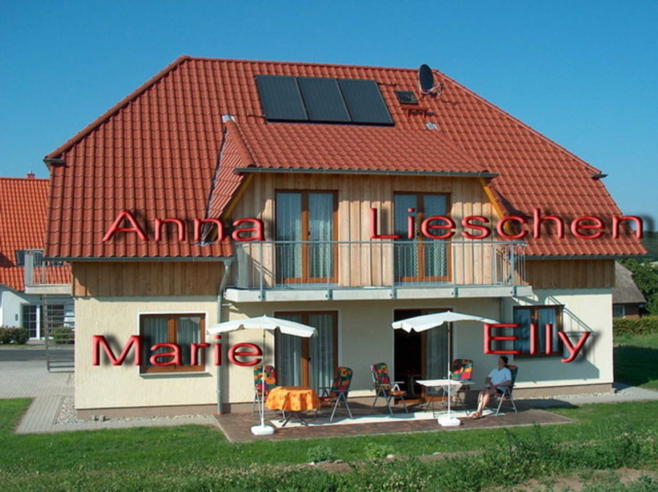 A two-story house with a red-tiled roof, a balcony, and a covered outdoor seating area with chairs and a table.