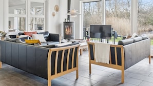 A modern living room with a large sofa, a wooden coffee table, and a TV. There is a fireplace and large windows offering a view of the outdoors. - Gorgeous home in Vejby with sauna (Vejby)