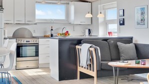 A modern kitchen with white cabinets, a built-in oven, a dining area with a gray sofa and a small wooden table, and a view of the sky through the window. - 2 bedroom amazing home in Hjørring (Hjørring)