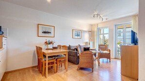 A spacious living area with a dining table, chairs, a sofa, and a wicker chair. There are framed pictures on the wall, a ceiling fan, and a view of the outside through large windows. - Villa Aquamarina Whg. 15, BALCONY, CHAIR - Villa Aquamarina, 1st FLOOR, partly. (Heringsdorf)