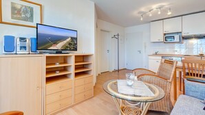A modern living room with a television, wooden furniture, a glass table, and a kitchen area with white cabinets and a microwave. - Villa Aquamarina Whg. 15, BALCONY, CHAIR - Villa Aquamarina, 1st FLOOR, partly. (Heringsdorf)