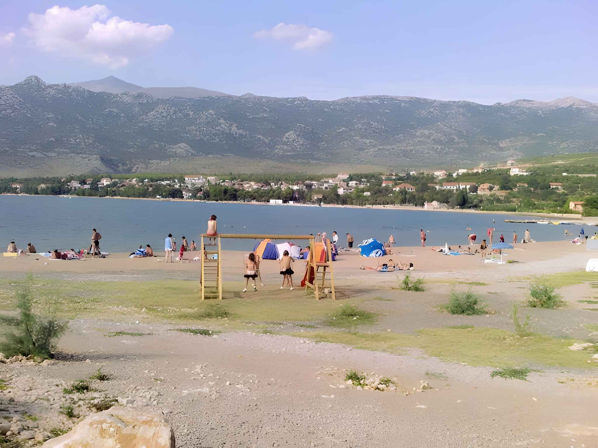 Une plage avec une aire de jeux, des gens qui prennent un bain de soleil et des maisons en arrière-plan.