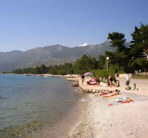 Ein Strand mit klarem Wasser, Kieselsteinen und Bergen im Hintergrund.