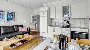 Ett modernt kök med vita skåp, en kaffemaskin och en spis. Det finns en matplats med ett bord och stolar, och ett vardagsrum med en svart soffa och konst på väggen.