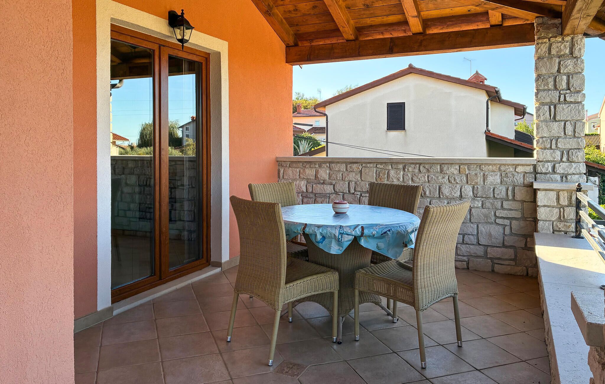 Un porche cubierto con una mesa redonda y sillas de mimbre, paredes de piedra y vistas a edificios cercanos.