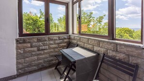 Een hoekbalkon met een tafel en bank, stenen muur en grote ramen met uitzicht op bomen en gebouwen.