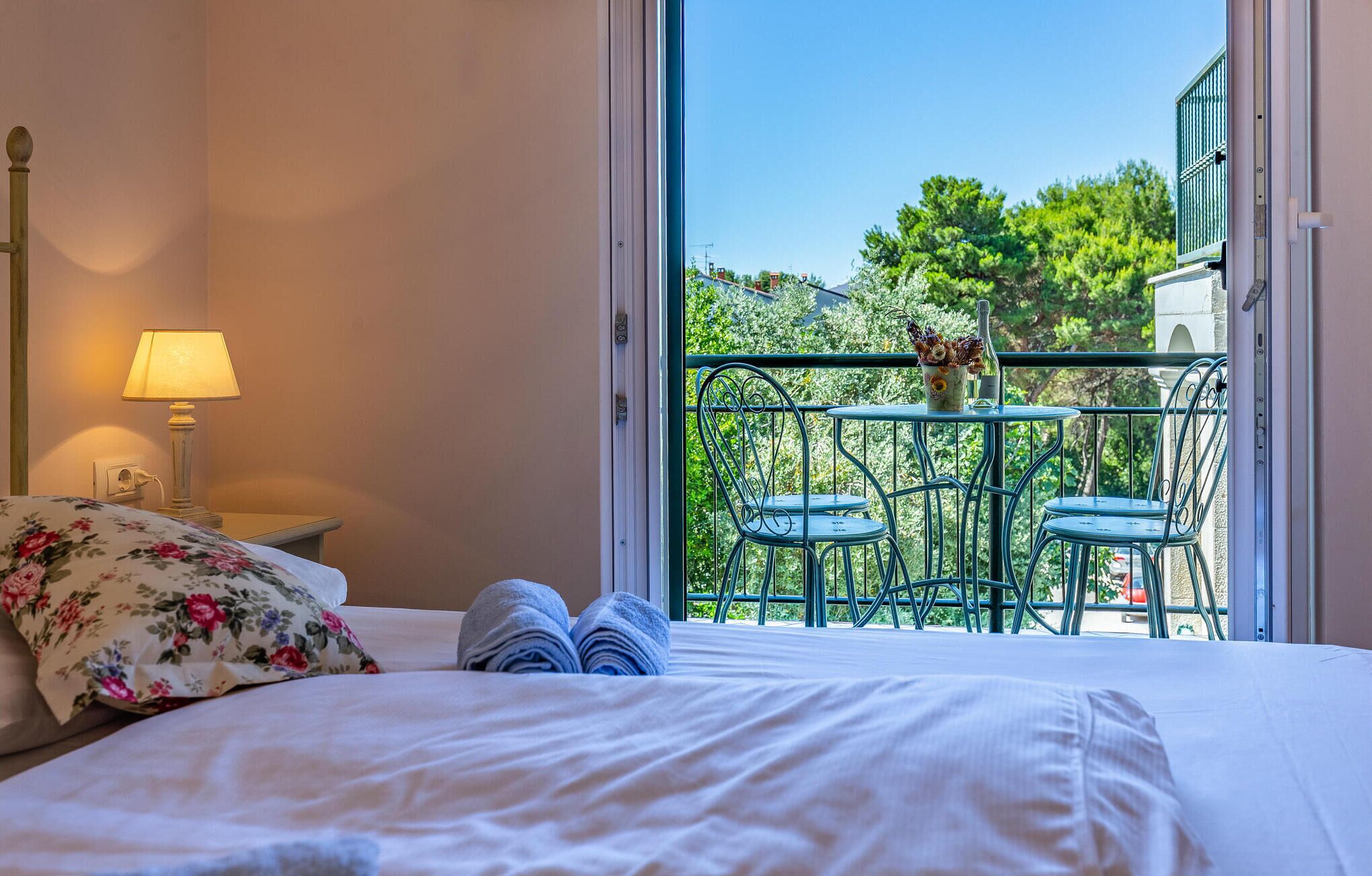 A hotel room with a bed, a nightstand with a lamp, and a balcony with two chairs and a table.