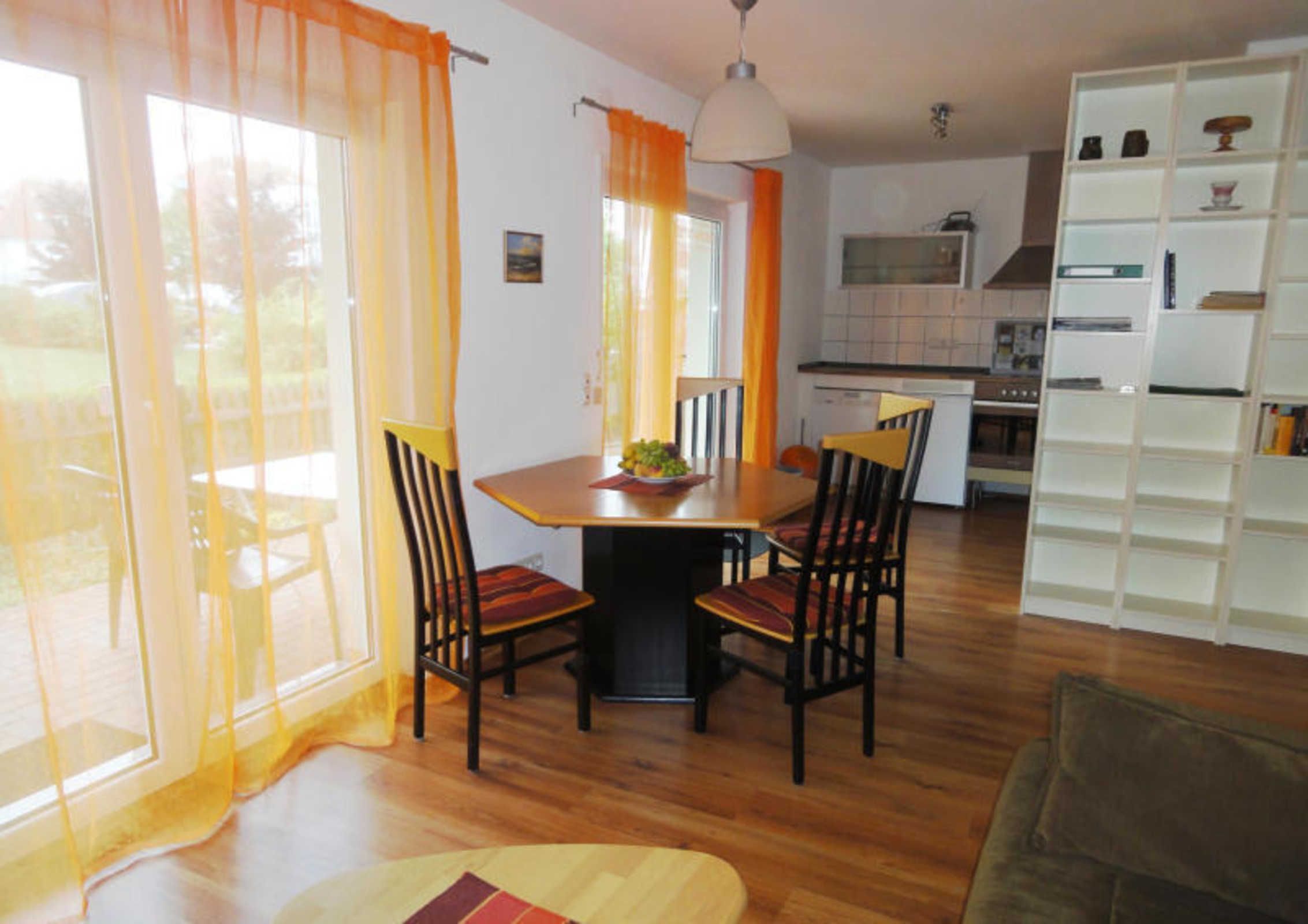A dining area with a wooden table and chairs, a kitchen in the background, and a white shelving unit.