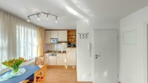 Une cuisine compacte avec des meubles blancs, un four à micro-ondes et un évier. On y trouve également un coin repas avec une table et des chaises, ainsi qu’une porte donnant sur une autre pièce.