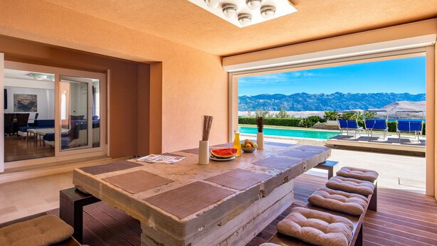 A modern room with a large window, a dining table with a view of mountains, and a pool area with lounge chairs. - 5 bedroom awesome home in Pag (Pag)