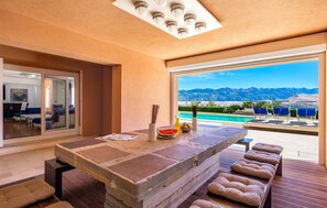 Une chambre moderne dotée d’une grande fenêtre, d’une table à manger offrant une vue sur les montagnes et d’un espace piscine avec des chaises longues.