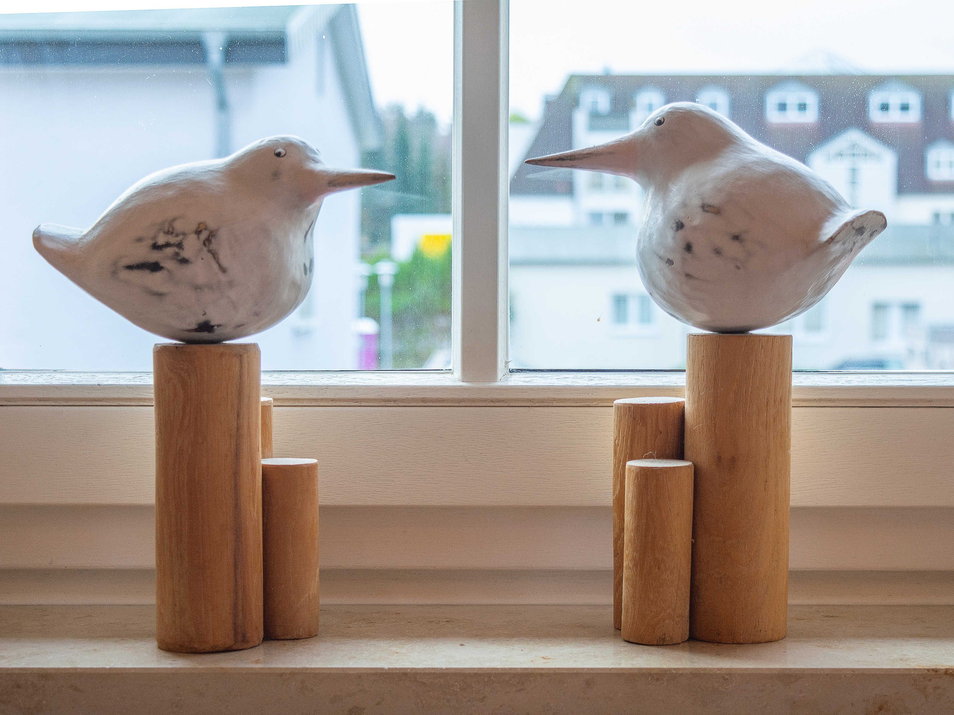 Dos pájaros de cerámica sobre pedestales de madera junto a una ventana.