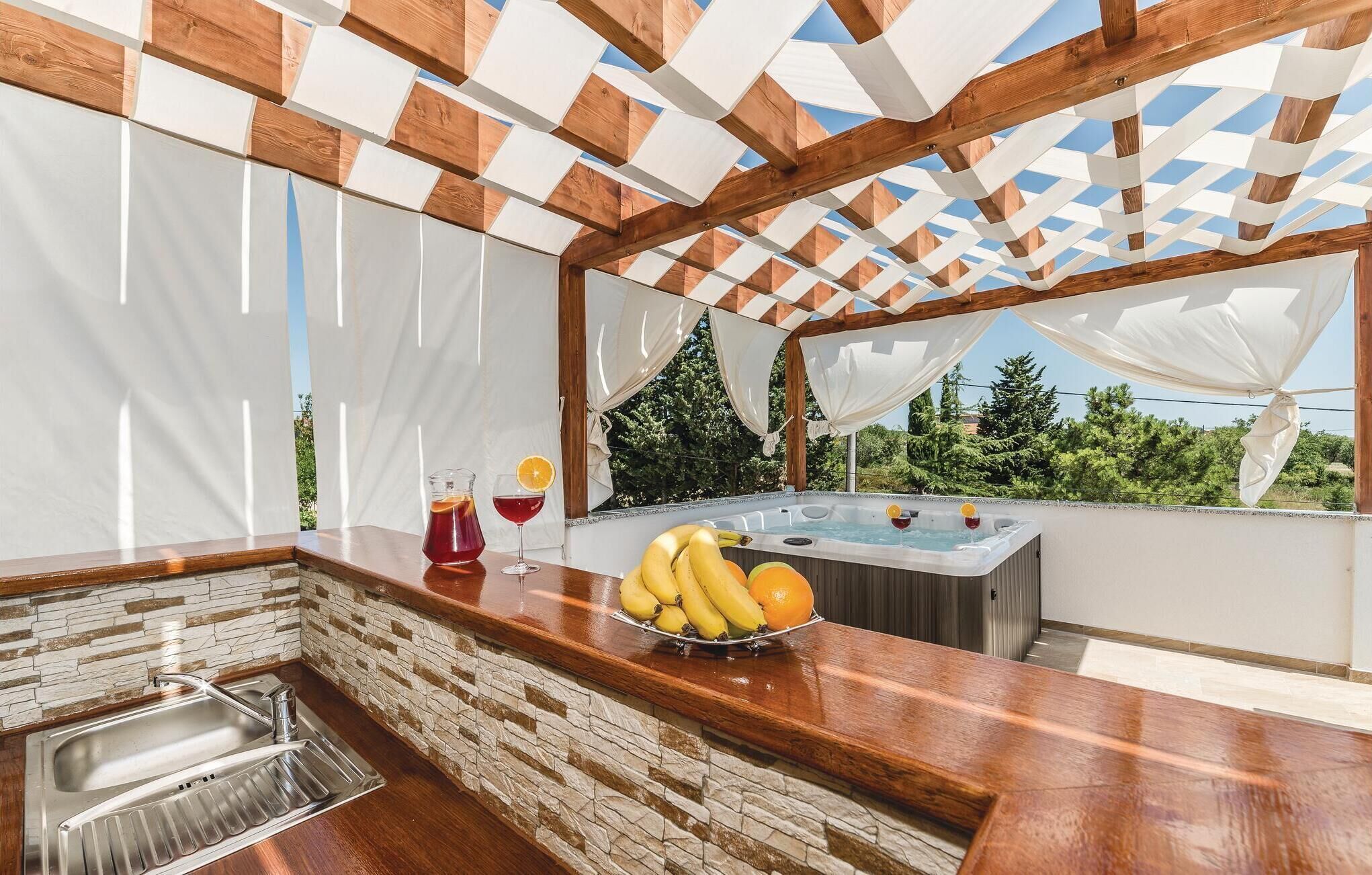 An outdoor kitchen area with a stone backsplash, a wooden countertop, a hot tub, and a view of the surrounding greenery.