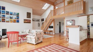 Amplia sala de estar con escalera de madera, comedor con mesa de vidrio y cocina con armarios blancos.