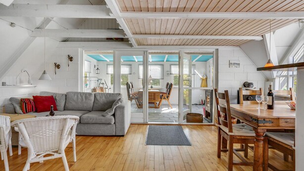 Un loft spacieux avec de hauts plafonds, un plancher en bois, un canapé gris, des chaises en osier, une table à manger avec des chaises, et une vue sur un espace extérieur grâce à de grandes portes vitrées.