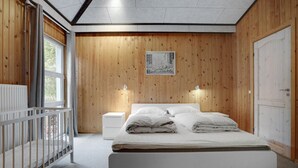 A bedroom with a wooden paneled wall, a bed with white bedding, a nightstand, a crib, and a window with curtains. - Nice home in Ebeltoft with kitchen (Ebeltoft)