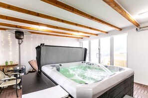 A modern bathroom with a large bathtub, wooden beams, a floor lamp, and a view of the outdoors. - Holiday apartment with sauna, whirlpool, garden and playground (Hévíz)