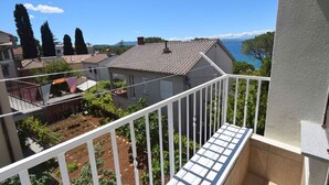 Un balcón con baranda blanca que da a un jardín y vistas a casas y al mar.