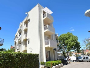 A multi-story residential building with balconies, a cross on the facade, and parked vehicles in the foreground.