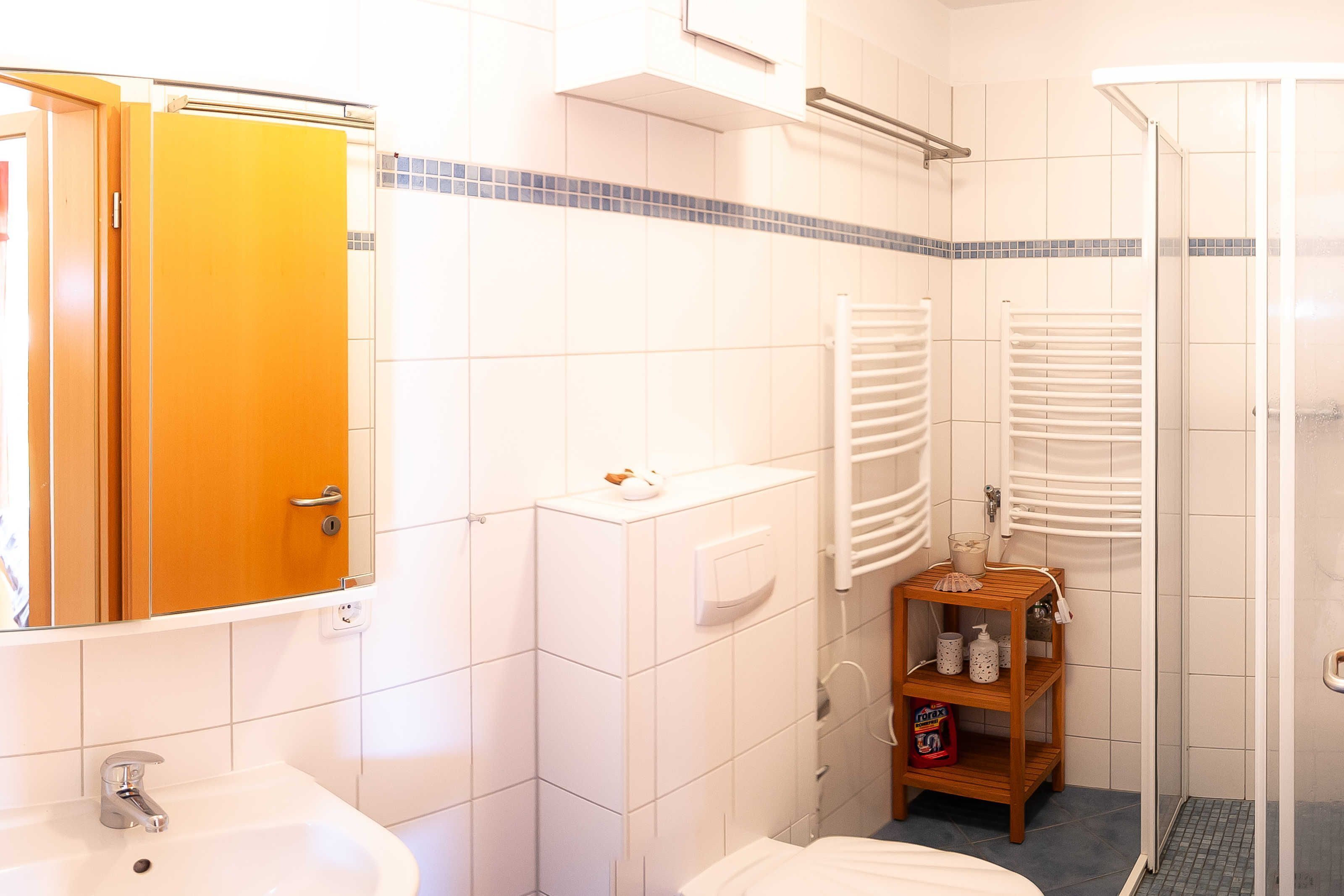 Casa de banho com lavatório, espelho, porta-toalhas, chuveiro e pequena prateleira com produtos de higiene pessoal.