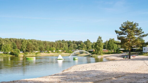 Vista para um lago com praia de areia, fileira de cabanas e área arborizada ao fundo.