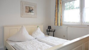 Uma cama bem arrumada com roupa de cama e almofadas brancas, um quadro na parede, uma luminária sobre uma mesinha de cabeceira e uma janela com cortinas.