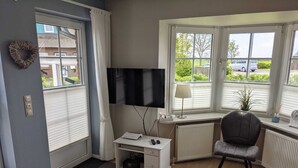 Ein modernes Wohnzimmer mit Fernseher, einem weißen Schrank, einem grauen Sessel und Blick durch große Fenster nach draußen.