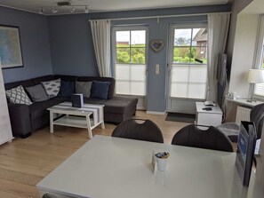 A modern living room with a grey sofa, white coffee table, and a dining table with chairs. There are two windows with curtains, a flat-screen TV, and a lamp on a bedside table.