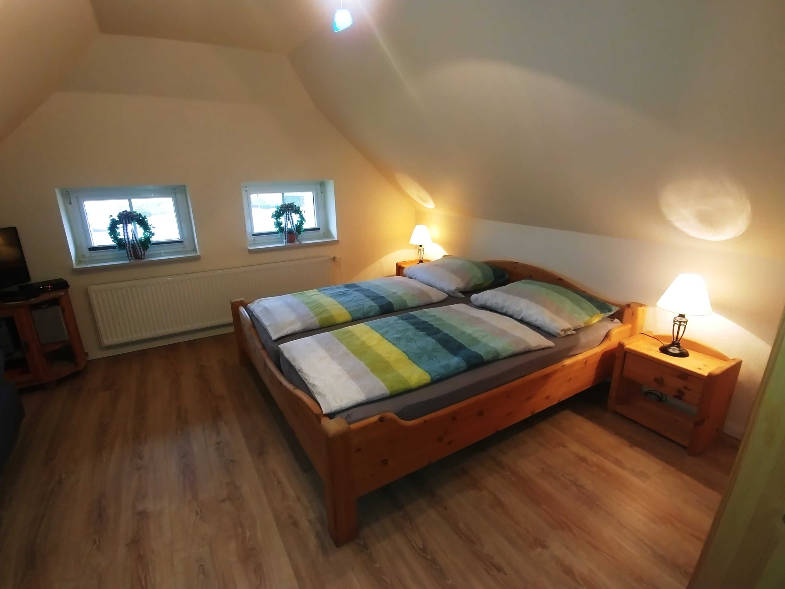 A bedroom with a wooden bed, two bedside tables with lamps, a radiator, and two windows with wreaths.