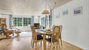 Zona pranzo con tavolo e sedie in legno, ampie finestre e quadri appesi al muro.