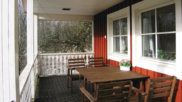 Eine überdachte Veranda mit Holztischen und -stühlen, großen Fenstern und Blick auf Bäume.