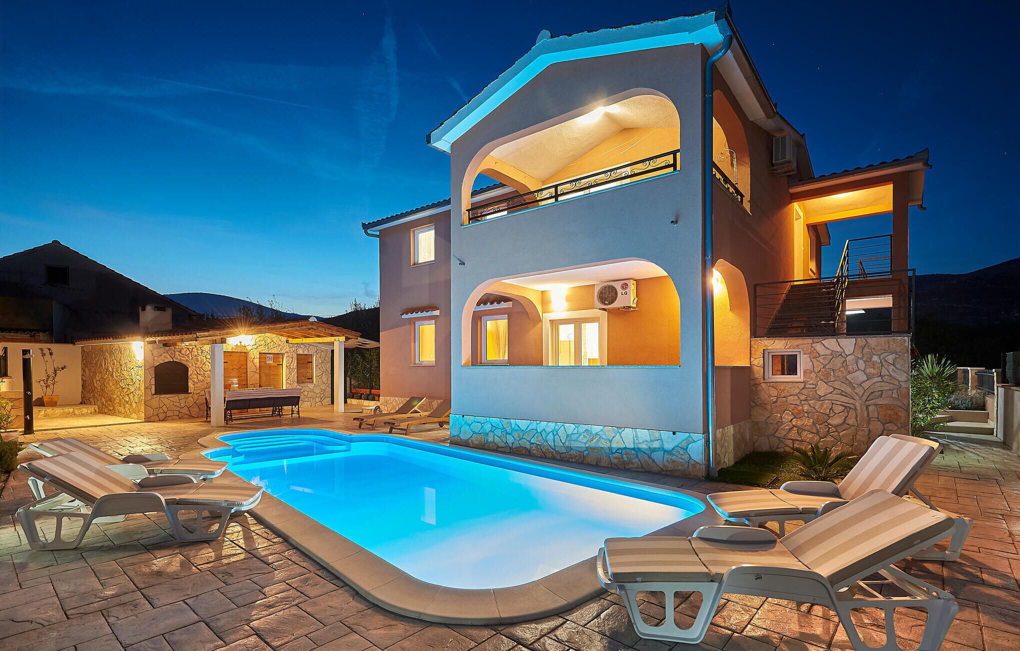Ein zweistöckiges Haus mit Swimmingpool, Liegestühlen und einer Fassade aus Naturstein.