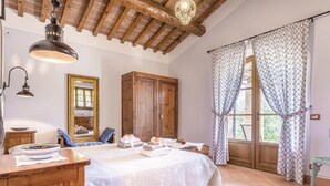 Un dormitorio con techo de madera, un armario grande, una cama con ropa de cama blanca, una mesita de noche, un espejo y una ventana con cortinas estampadas.