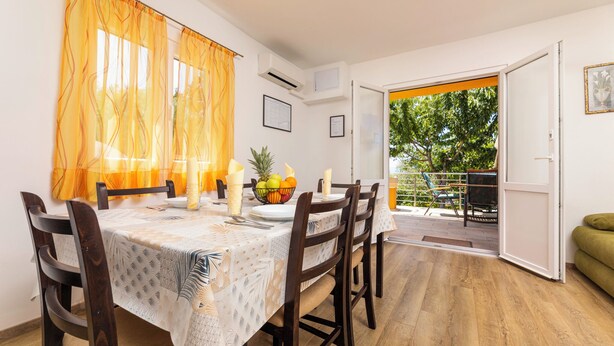 Zona pranzo con tavolo imbandito per quattro persone, una ciotola di frutta e vista sull'esterno.