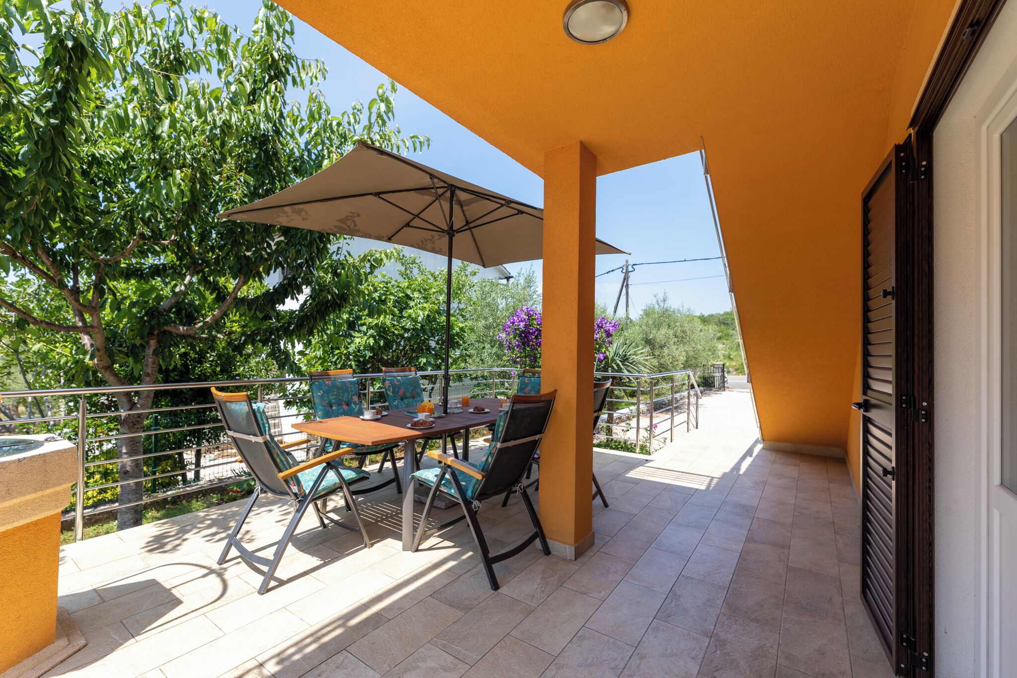 Um pátio externo com mesa e cadeiras, um grande guarda-sol e vista para árvores e flores roxas.