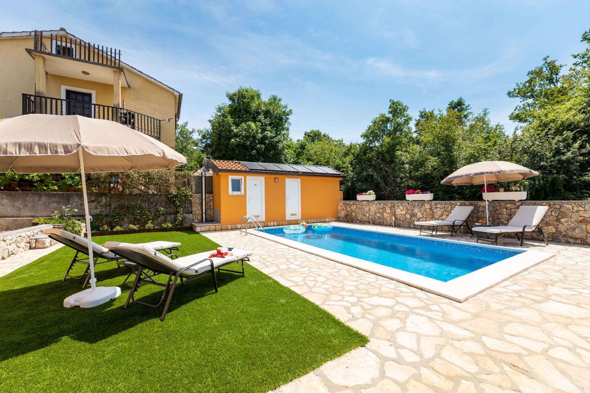 Une piscine entourée de chaises longues et de parasols, elle-même bordée d’une terrasse en pierre et d’un petit bâtiment.