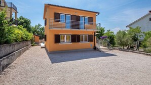 Edificio a due piani con un balcone, vialetto in ghiaia e vegetazione circostante.