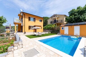 Una casa moderna de dos pisos con piscina, un patio con sillones de descanso y un jardín.