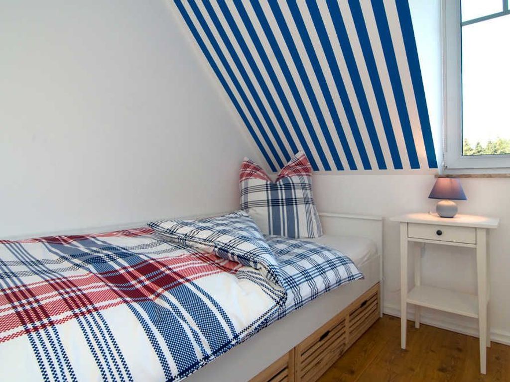 A bedroom with a sloped ceiling, a bed with a plaid bedspread, two pillows, a bedside table with a lamp, and a window.