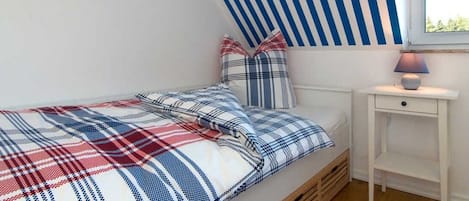A bedroom with a sloped ceiling, a bed with a plaid bedspread, two pillows, a bedside table with a lamp, and a window.