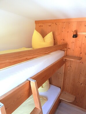 Ein Stockbett mit Holzrahmen und weißer Bettwäsche, eine Holzwand mit Ablagefläche und ein kleiner Holzschemel.